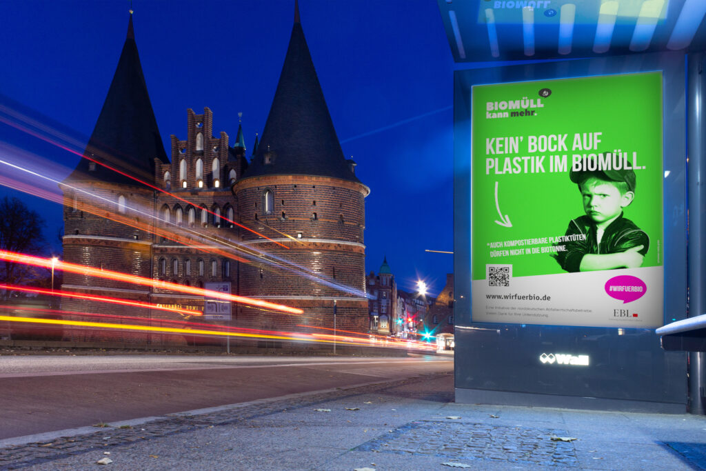 Langzeitbelichtung vom Verkehr vor dem Lübecker Holstentor im Hintergrund, im Vordergrund eine Bushaltestelle mit einem #wirfuerbio Kampagnenplakat.