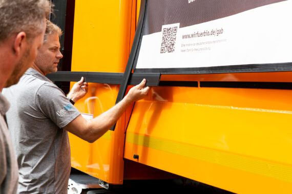 Thomas Pedersen von flexsign spannt einen Banner der Kampagne #wirfuerbio am orangefarbigen Müllfahrzeug ein.